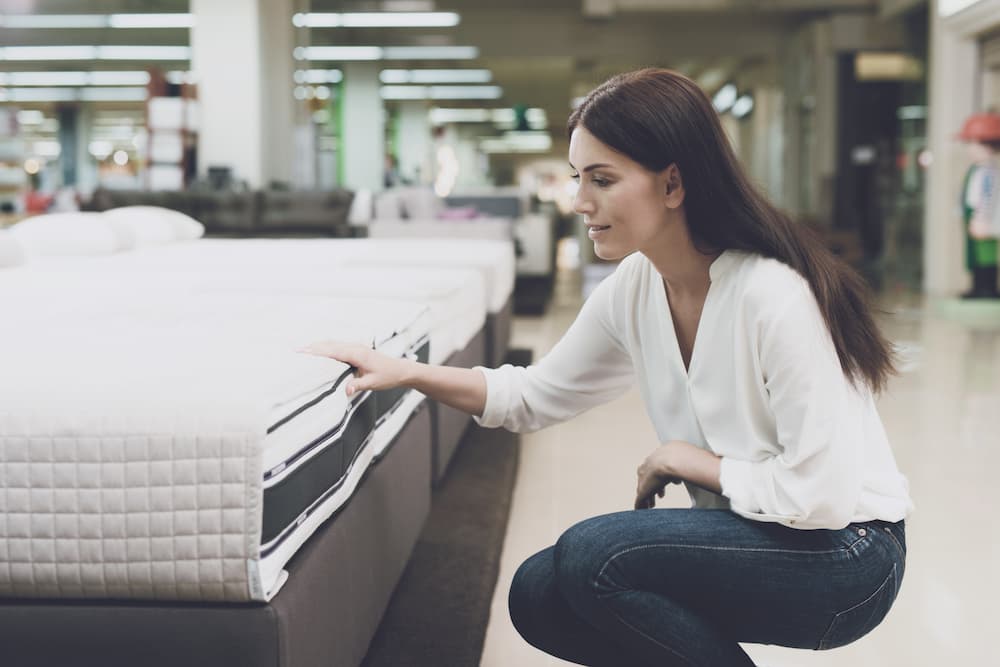 A woman in a white shirt and jeans in a mattress store. She examines the mattress she wants to buy.